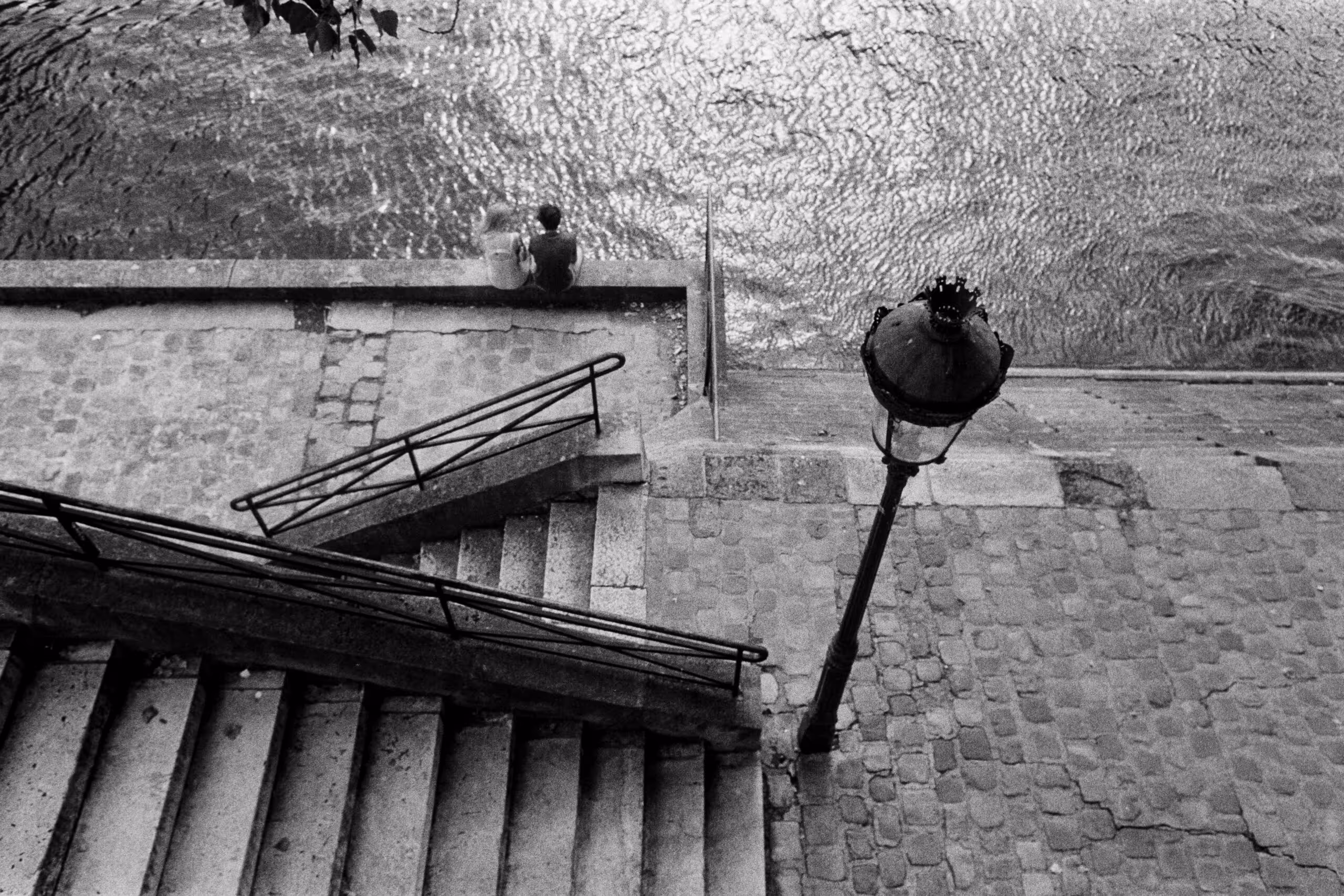 A Seine quay seen from the street above, stone steps and a lamppost in the foreground and a couple sitting alone at the water, shot on black and white film