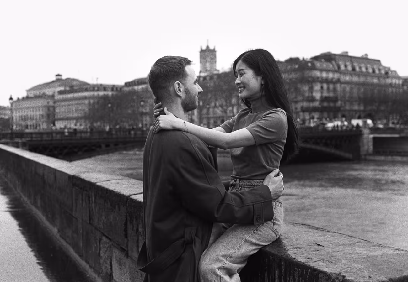 Couple on the stone wall of a Seine quay, Haussmann façades and a bridge behind them, shot on black and white film