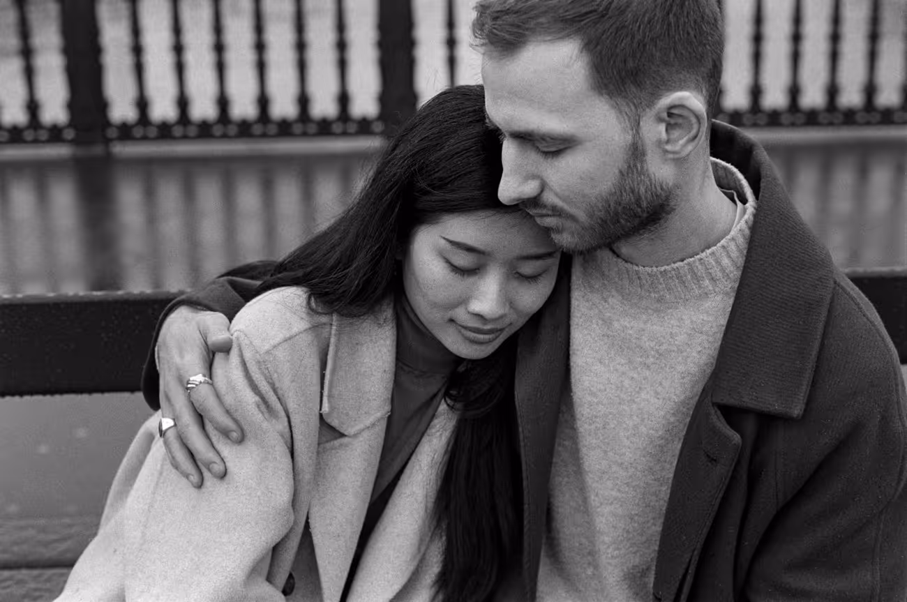 Man kissing his partner's forehead as she leans into his shoulder, both in winter coats, shot close on black and white film