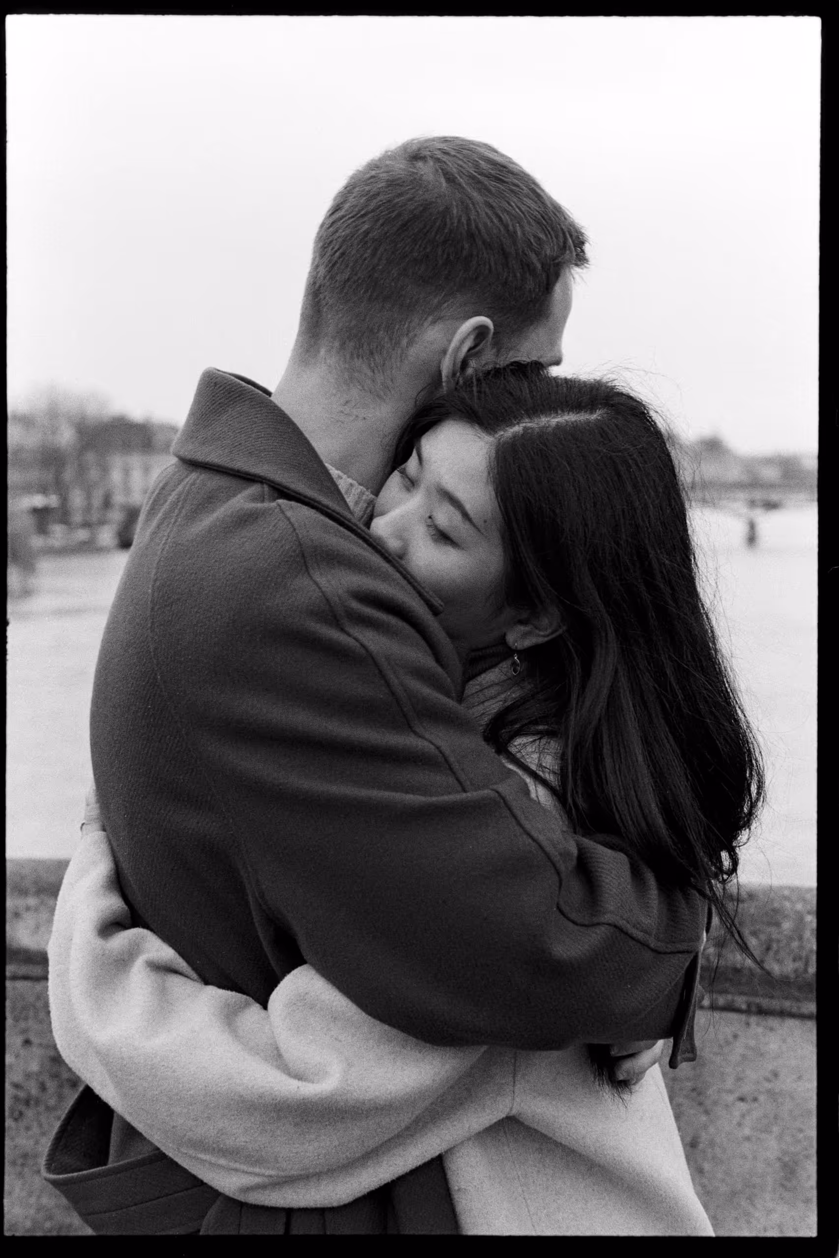 Couple holding each other tightly against a stone parapet in winter, the background blurred behind them, shot on black and white film
