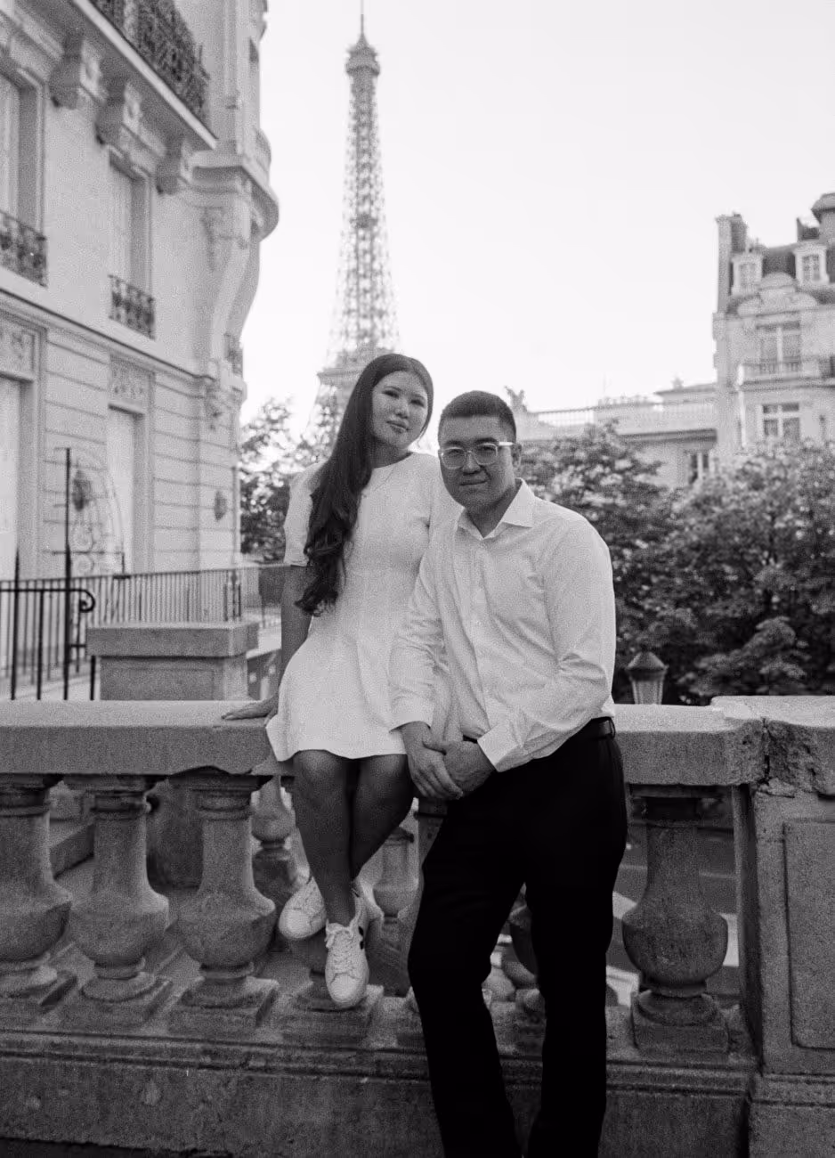 Couple sitting on the stone balustrade of the Avenue de Camoëns with the Eiffel Tower rising between the buildings behind them, shot on black and white film