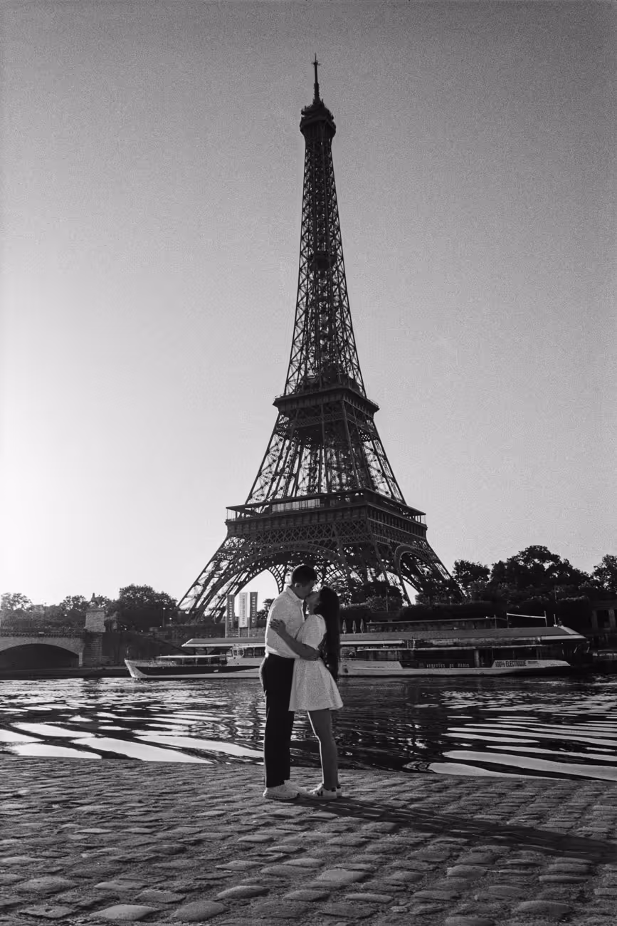 Couple embracing on the bank of the Seine at the foot of the Eiffel Tower, a river boat passing behind them, shot on black and white film