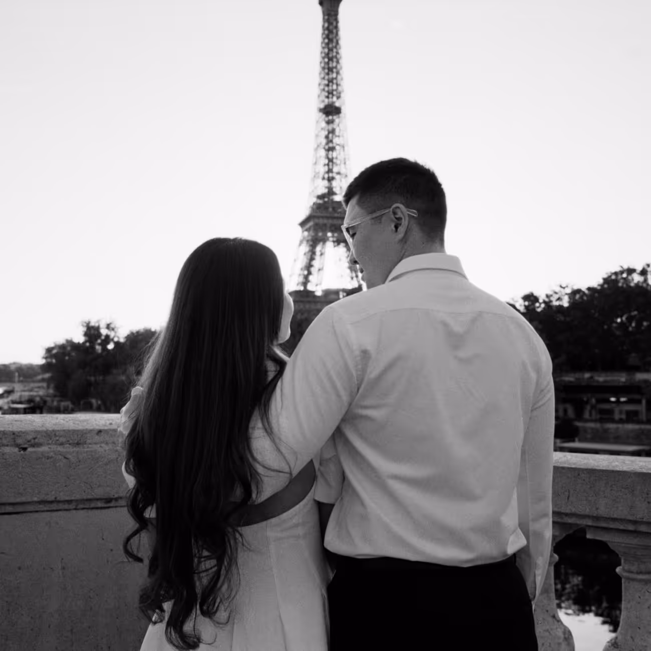 Couple photographed from behind at a stone parapet, looking out at the Eiffel Tower in the early evening light, shot on black and white film