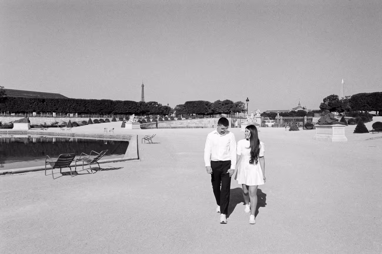 Couple walking hand in hand across the wide gravel of the Jardin des Tuileries, the basin and its green chairs beside them and the Eiffel Tower small on the skyline, shot on black and white film