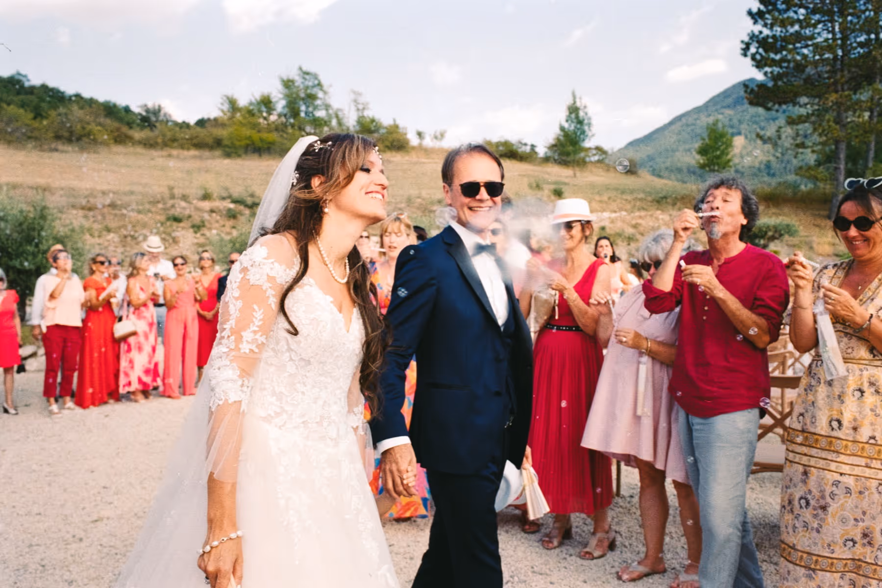 Bride and groom walking hand in hand through their guests, who blow bubbles, on a gravel path below the Provence hills at their Sisteron wedding