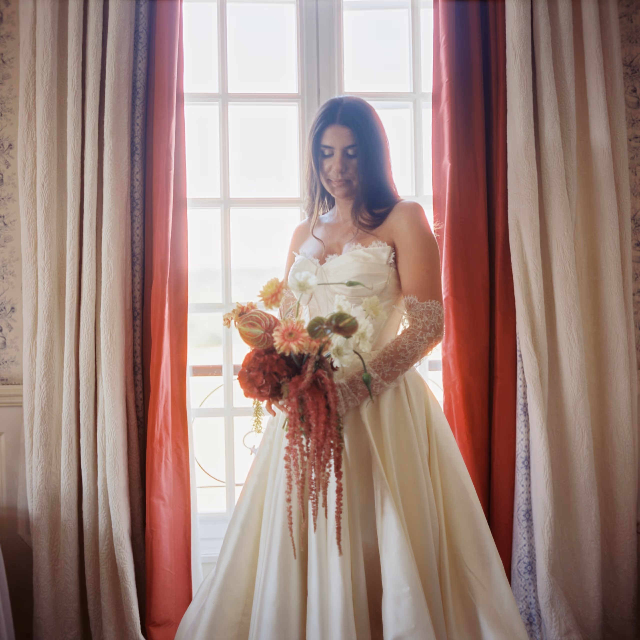 Bride holding her bouquet in the light of a tall window, framed by red curtains, provence wedding, south of france