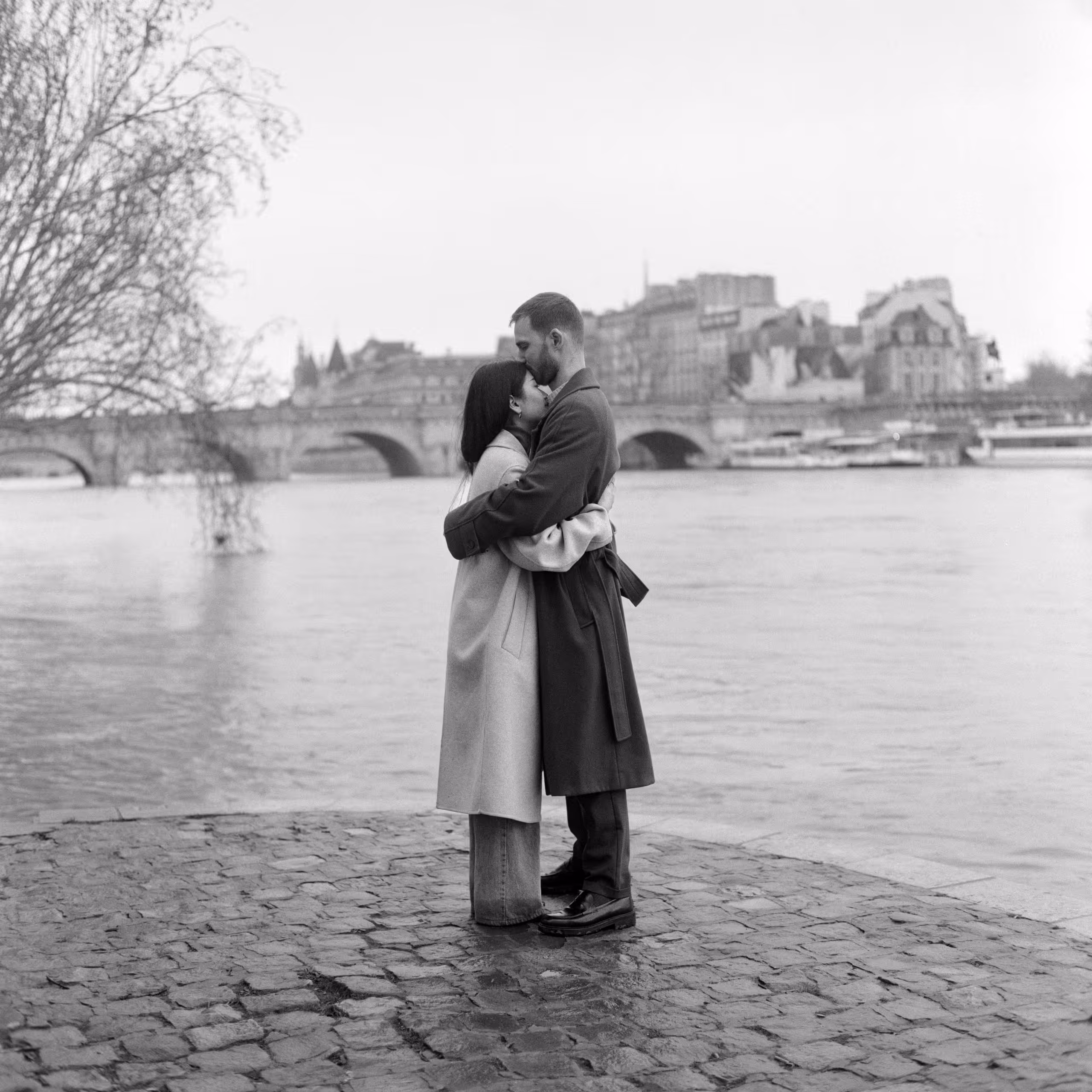 Couple embracing on the cobbled bank of the Seine with a Paris bridge behind them, shot on black and white film
