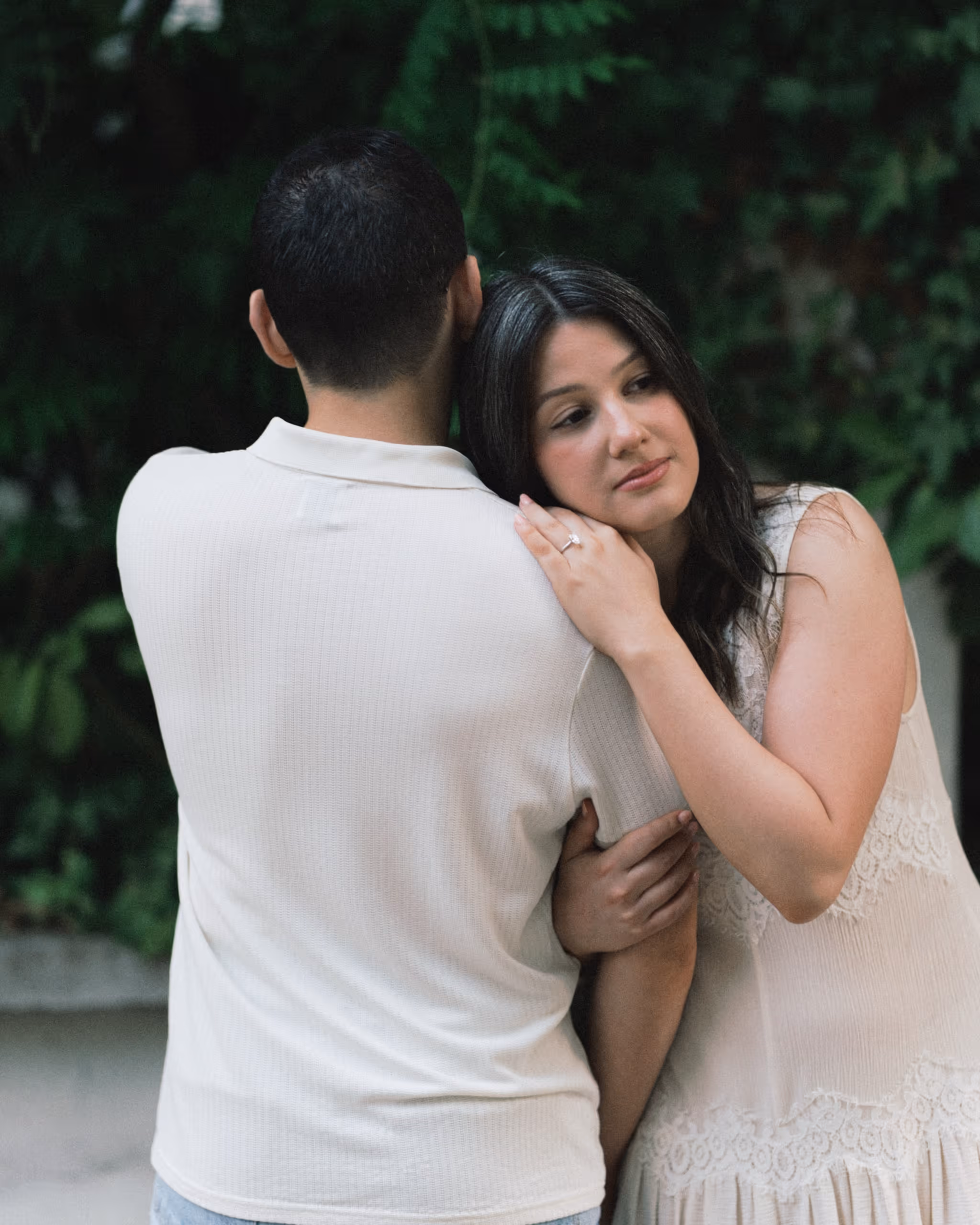 A woman resting her head on her partner's shoulder in a close embrace, her hand on his shoulder, in front of dense green foliage in Montmartre.