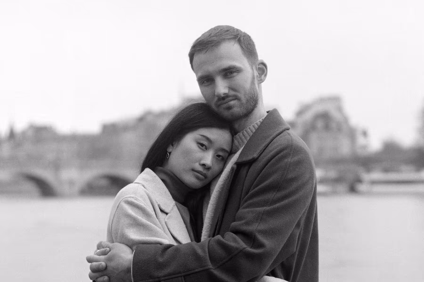 Black and white film portrait of a couple holding each other on the quais of the Seine, her head resting on his chest, with a bridge and the Louvre behind them