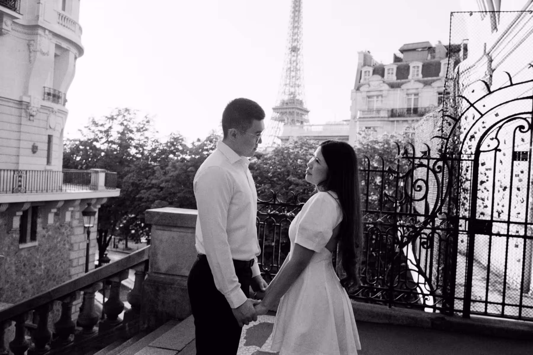 Black and white film photograph of a couple holding hands and looking at each other on a balcony at Avenue de Camoens, the Eiffel Tower rising behind them