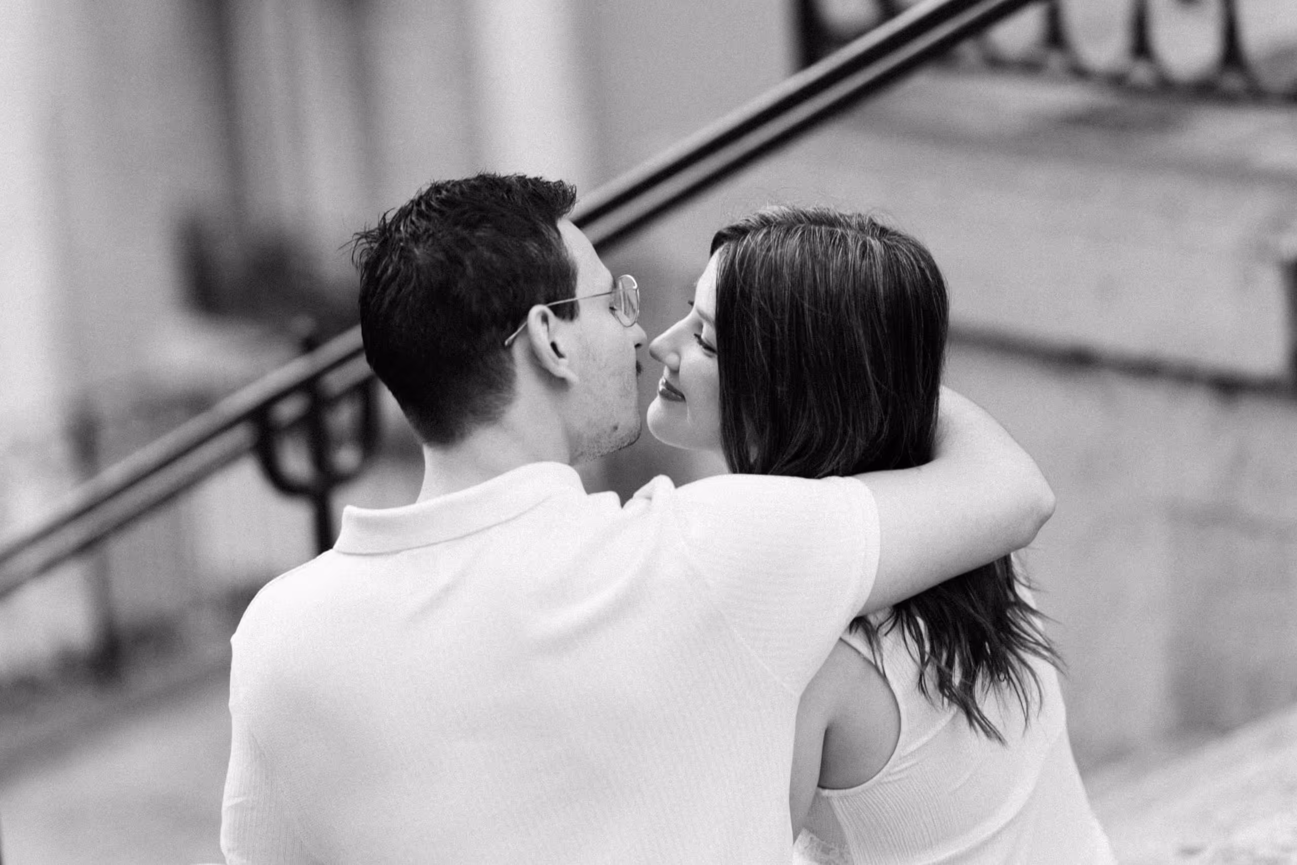 A black-and-white photograph of a couple sitting on a Montmartre stairway, her arm around his neck, their faces close as she smiles.