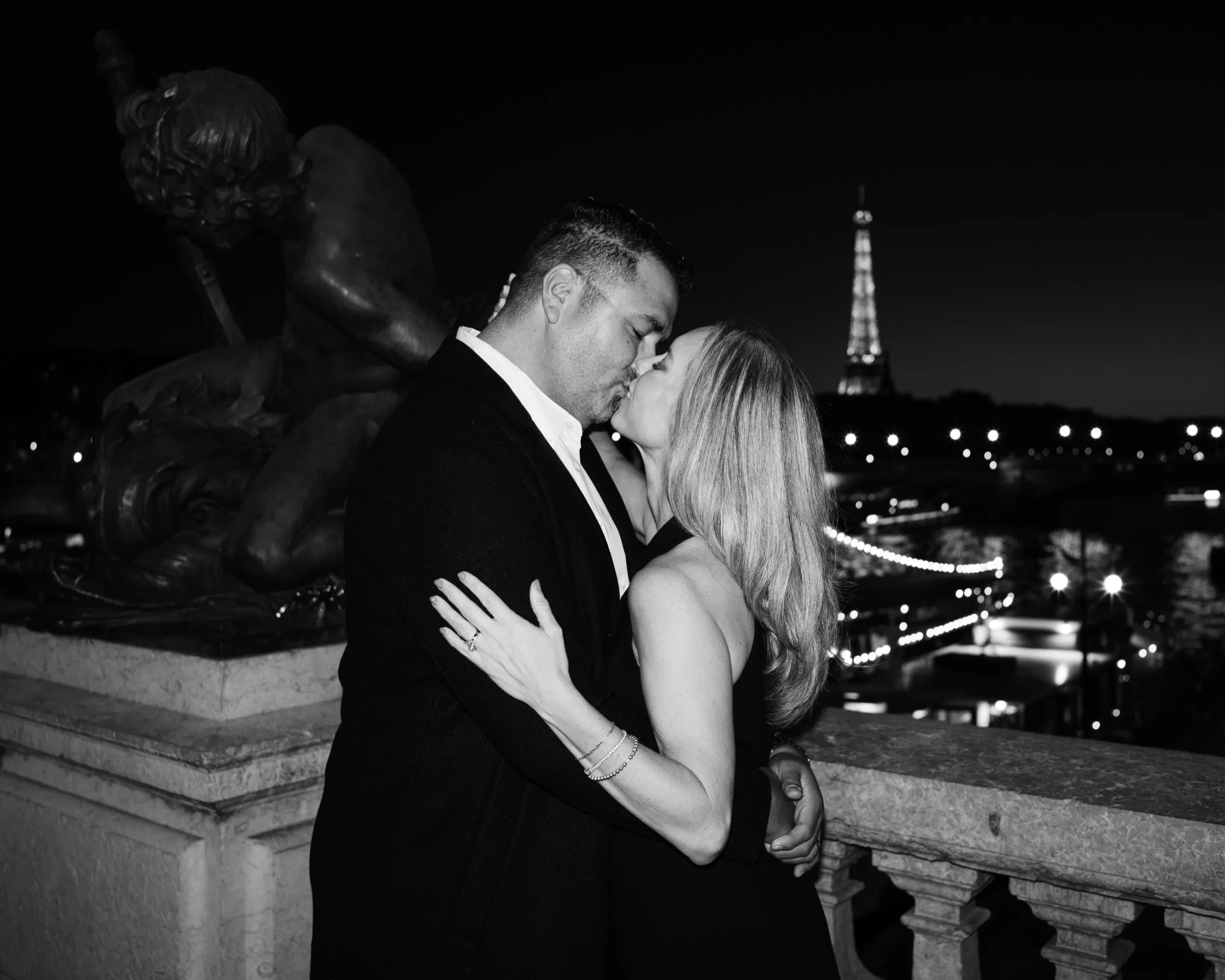 Couple kissing beside a bronze cherub on Pont Alexandre III at night, the lit Eiffel Tower behind them, in black and white