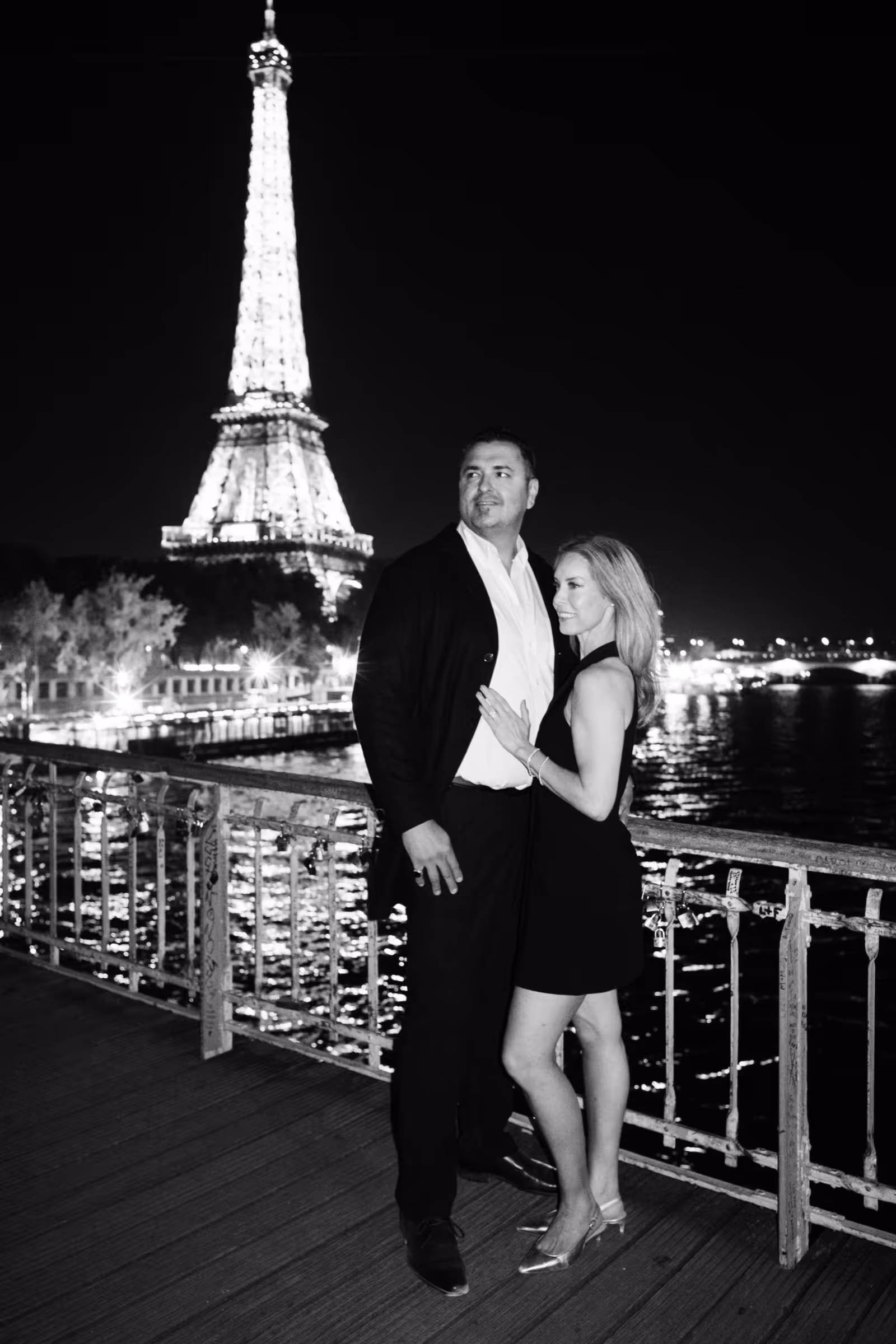 A couple standing close on the wooden deck of Passerelle Debilly at night, beside a railing covered in padlocks, with the lit Eiffel Tower rising behind them, in black and white.