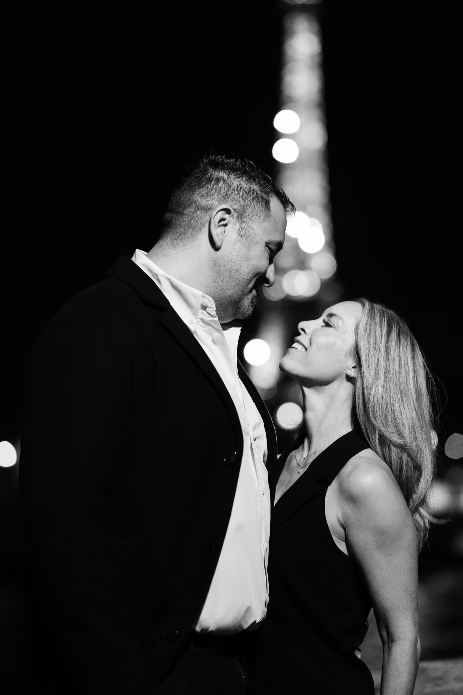 Close portrait of a couple smiling at each other at night, the lights of the Eiffel Tower blurred into soft points behind them, in black and white.