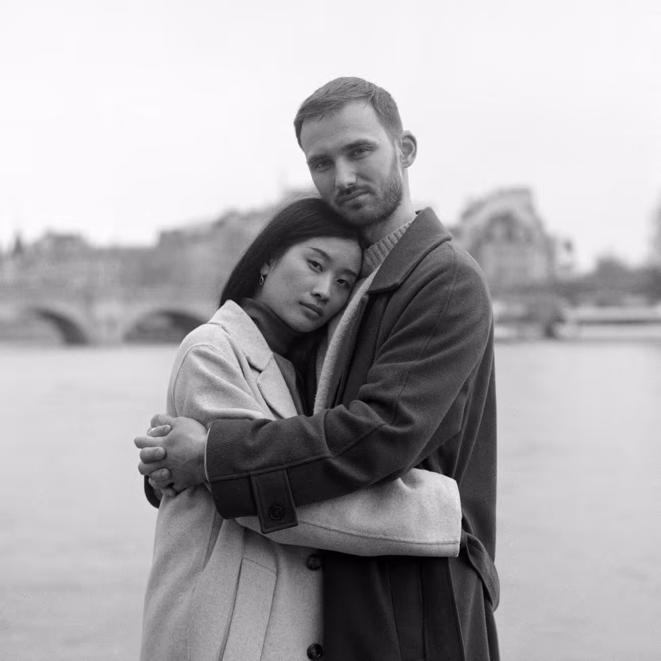 Suzanne and Titouan embracing beside the Seine in light and dark coats over knitwear, photographed in black and white.