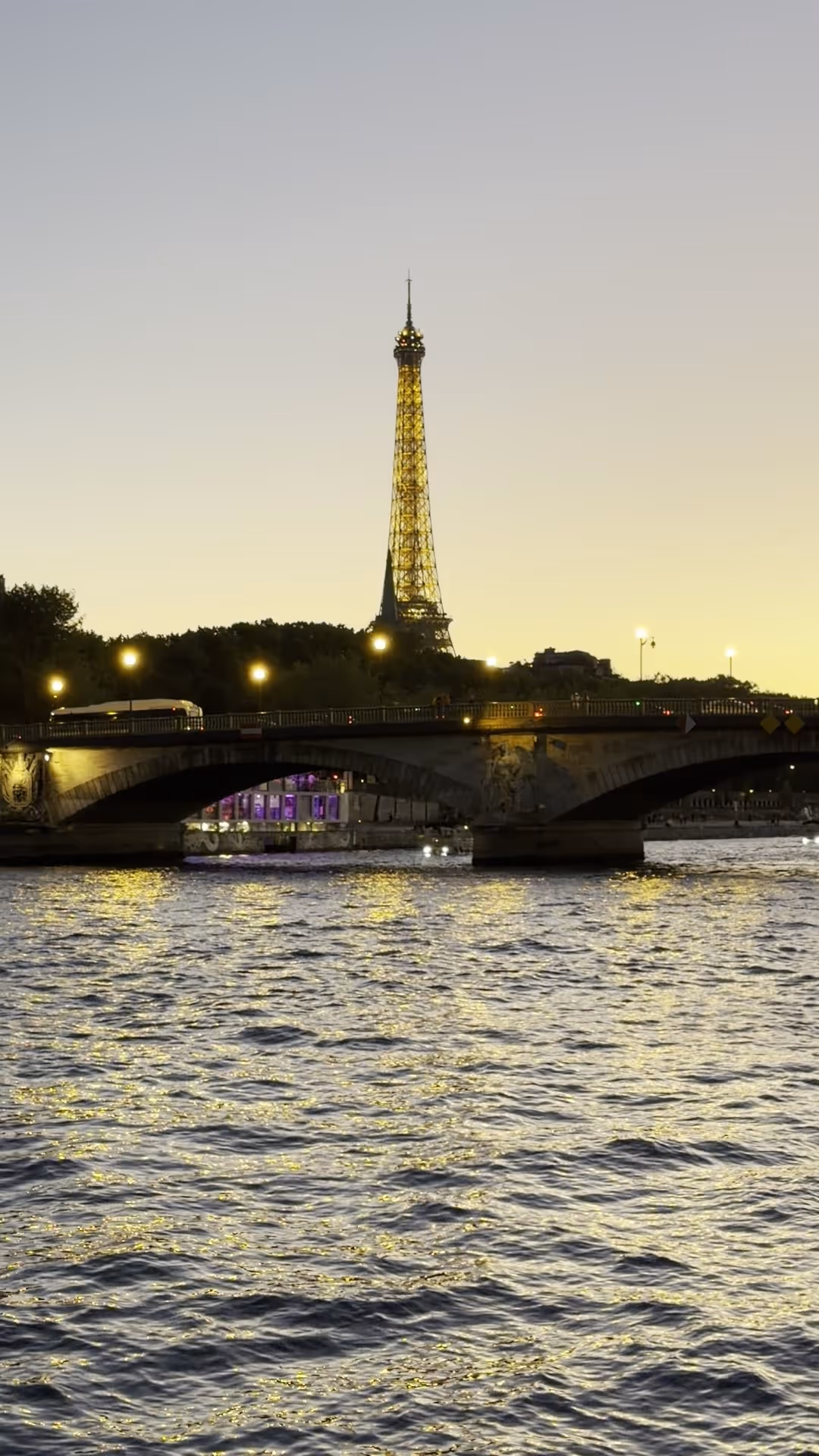 The Eiffel Tower lit in gold against a pale evening sky, seen down the Seine beyond a stone bridge, with the lights reflecting on the water.
