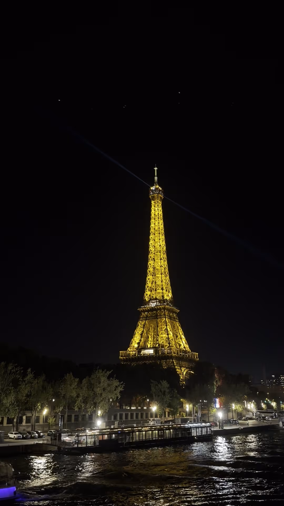 The Eiffel Tower lit in gold against a black sky, seen across the Seine at night, with the lights of the quays reflected in the water.