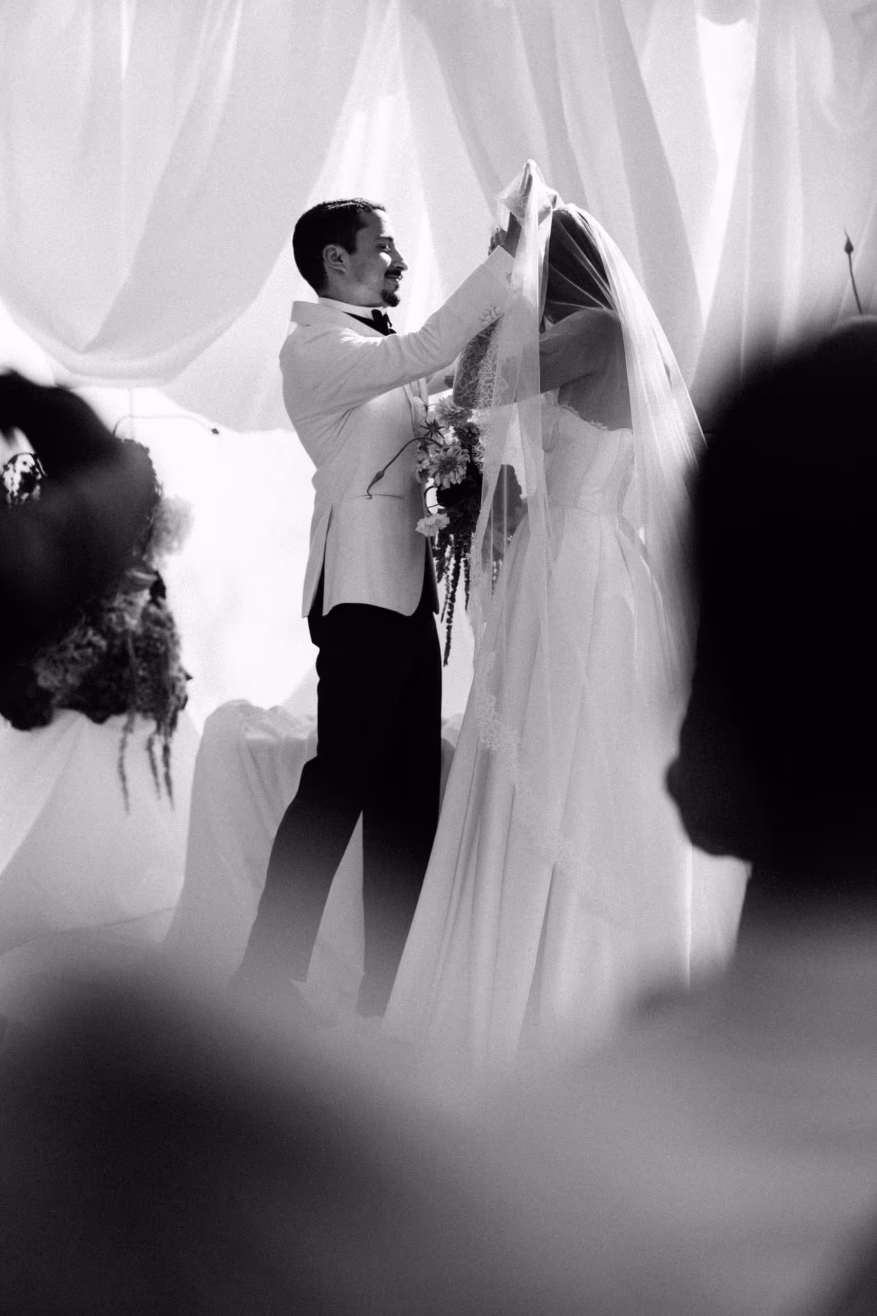 Groom lifting his bride's veil during the ceremony, seen between the guests