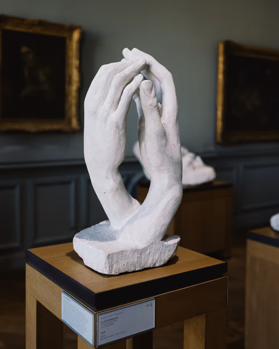 White marble sculpture of two hands on a wooden display stand in a Musée Rodin room.