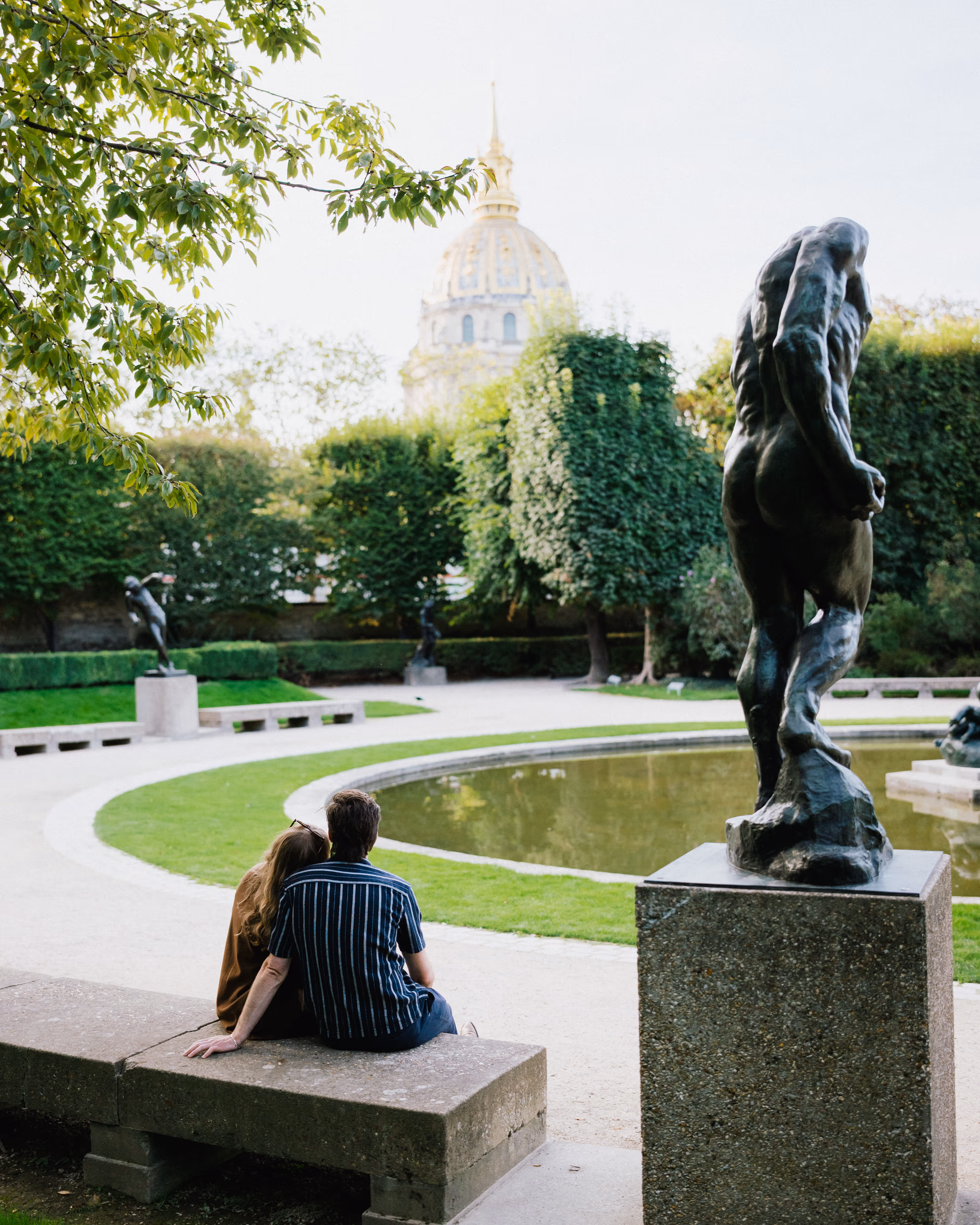 Couple seen from behind on a stone bench beside the Musée Rodin garden pond, with a bronze sculpture and the Invalides dome beyond