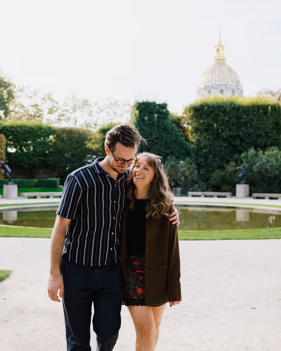 Paige and Reinhardt walking close together in front of the circular garden pond, with the Invalides dome above the hedge.