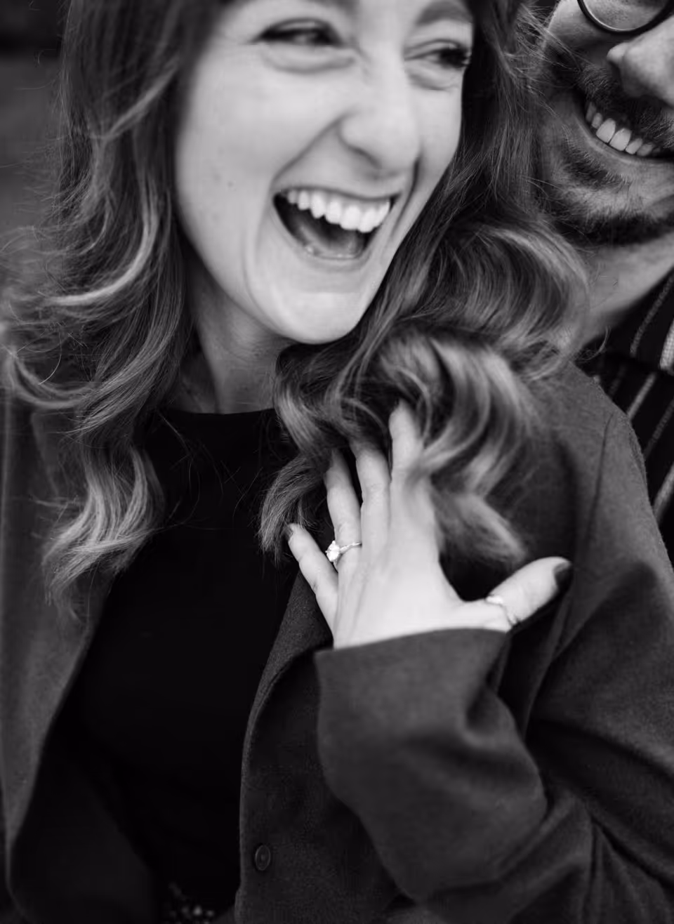 Black-and-white close portrait of Paige laughing beside Reinhardt, her left hand and engagement ring held near her chest.