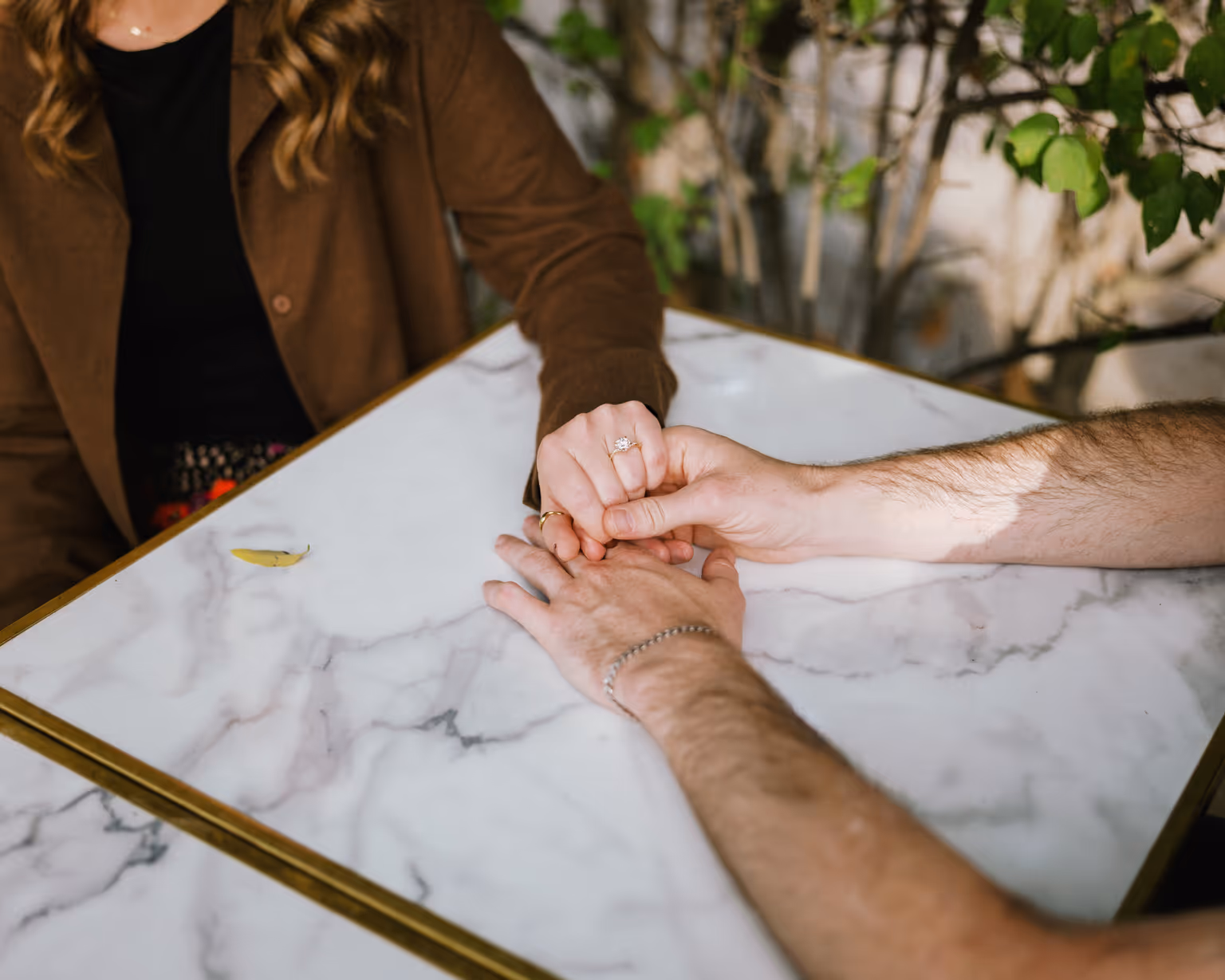 Paige and Reinhardt holding hands across a marble-topped table, with Paige's ring visible.