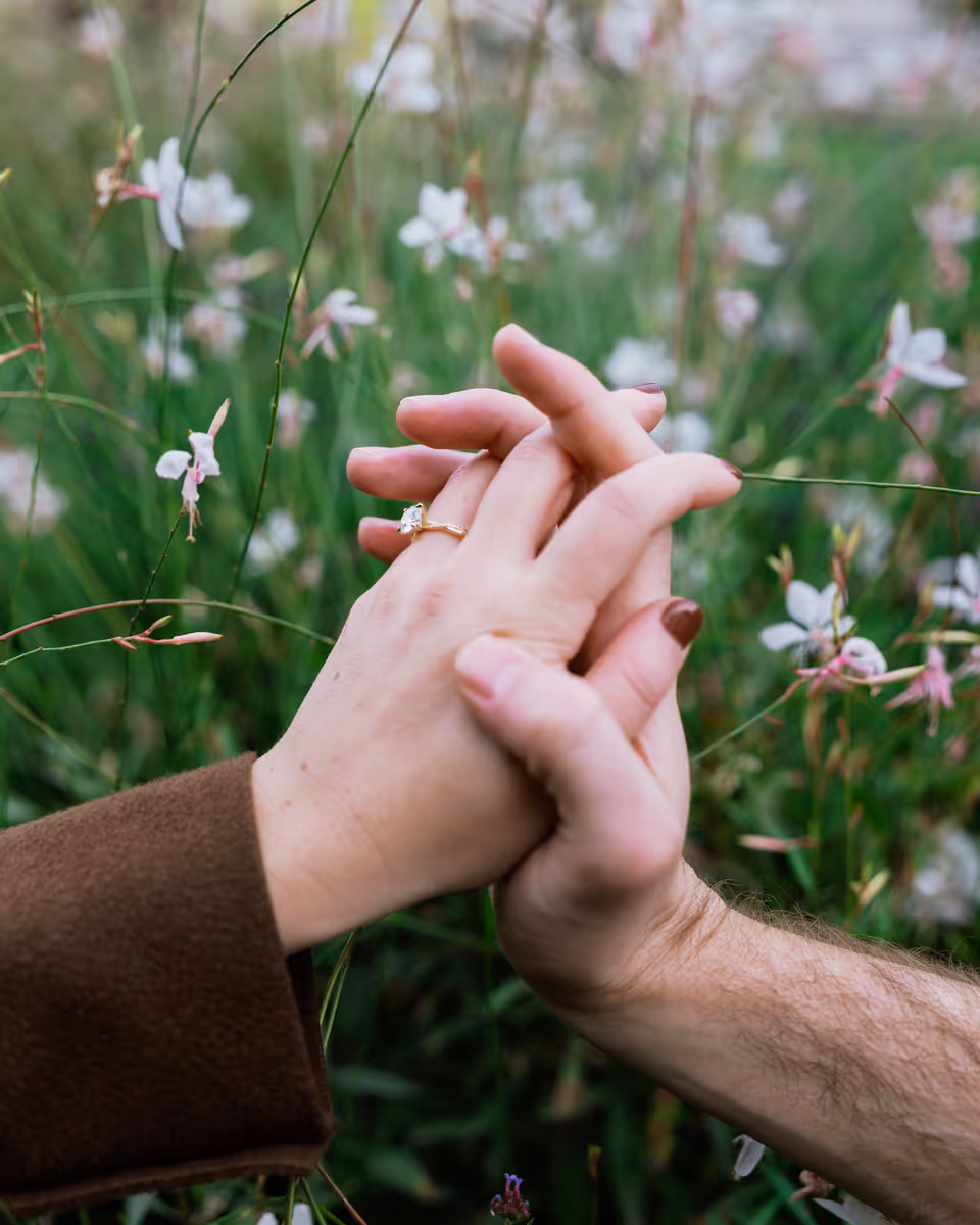Close view of Paige and Reinhardt's linked fingers in front of green planting and small pink flowers.