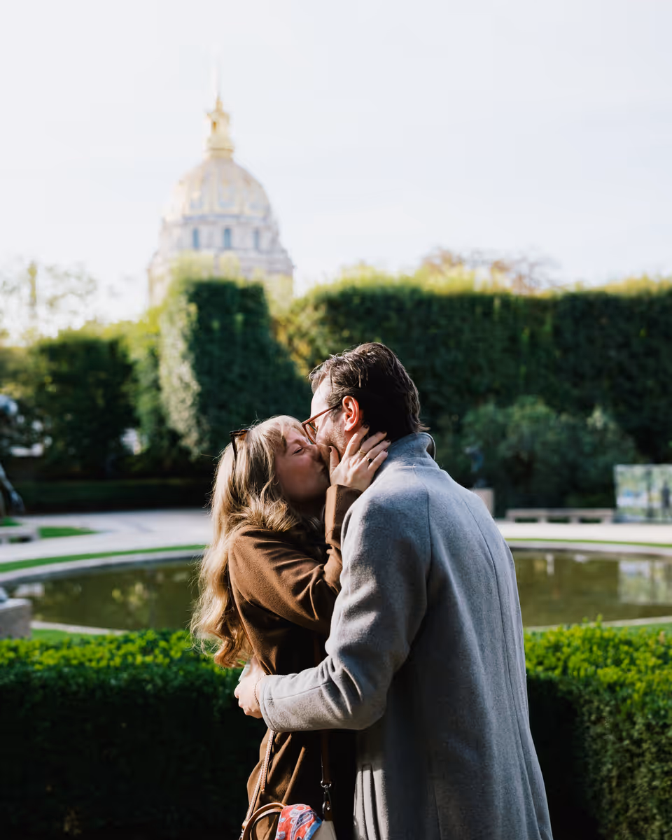 Paige and Reinhardt kissing in the Musée Rodin garden, with the Invalides dome behind them.