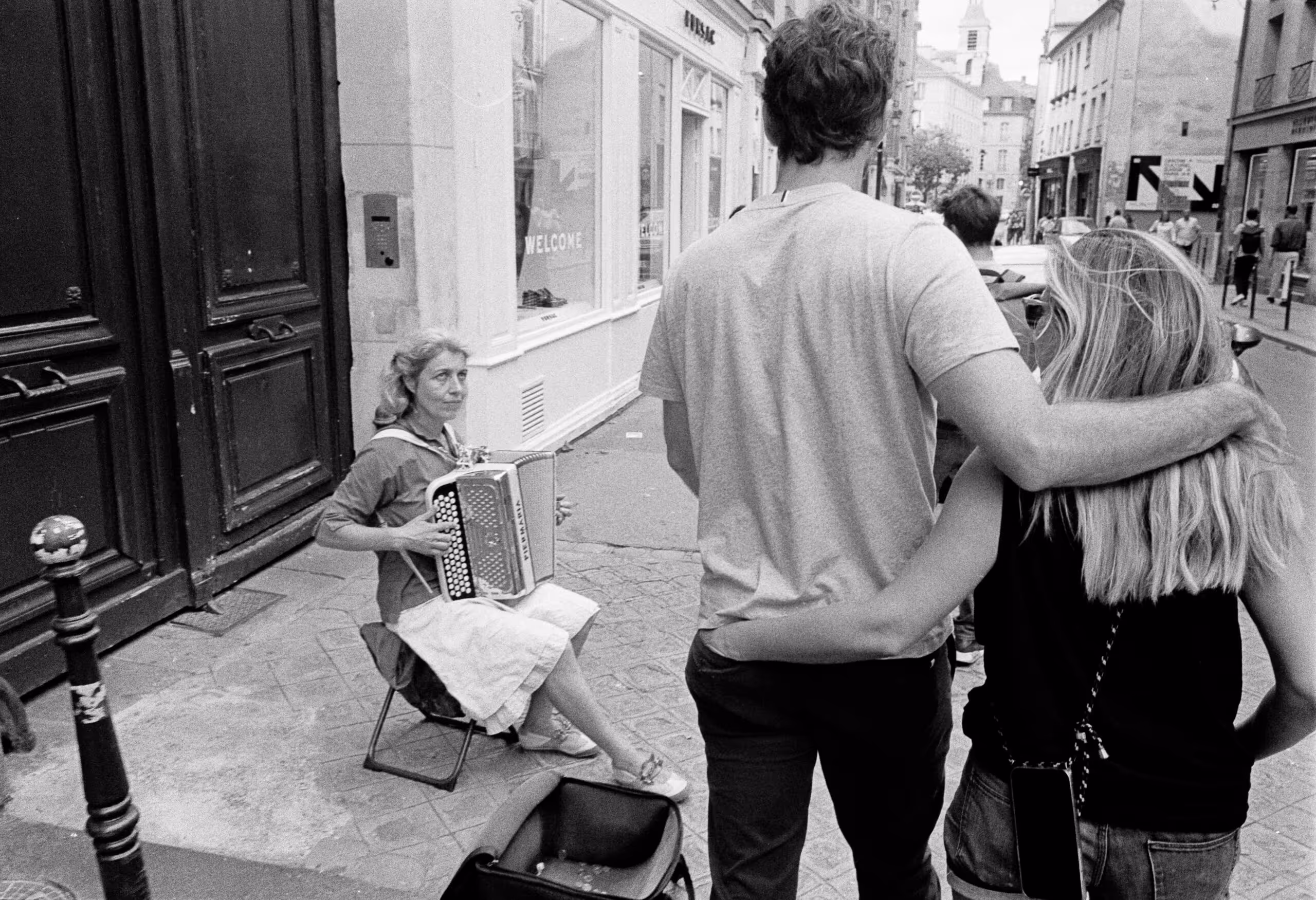 Couple walking arm in arm down a Paris street past a woman sitting playing the accordion, shot on black and white film